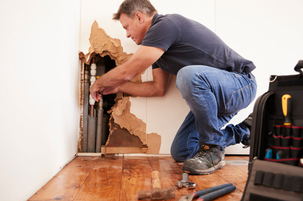 Person in the process of water mitigation, repairing pipes behind damaged drywall with tools scattered on the floor.